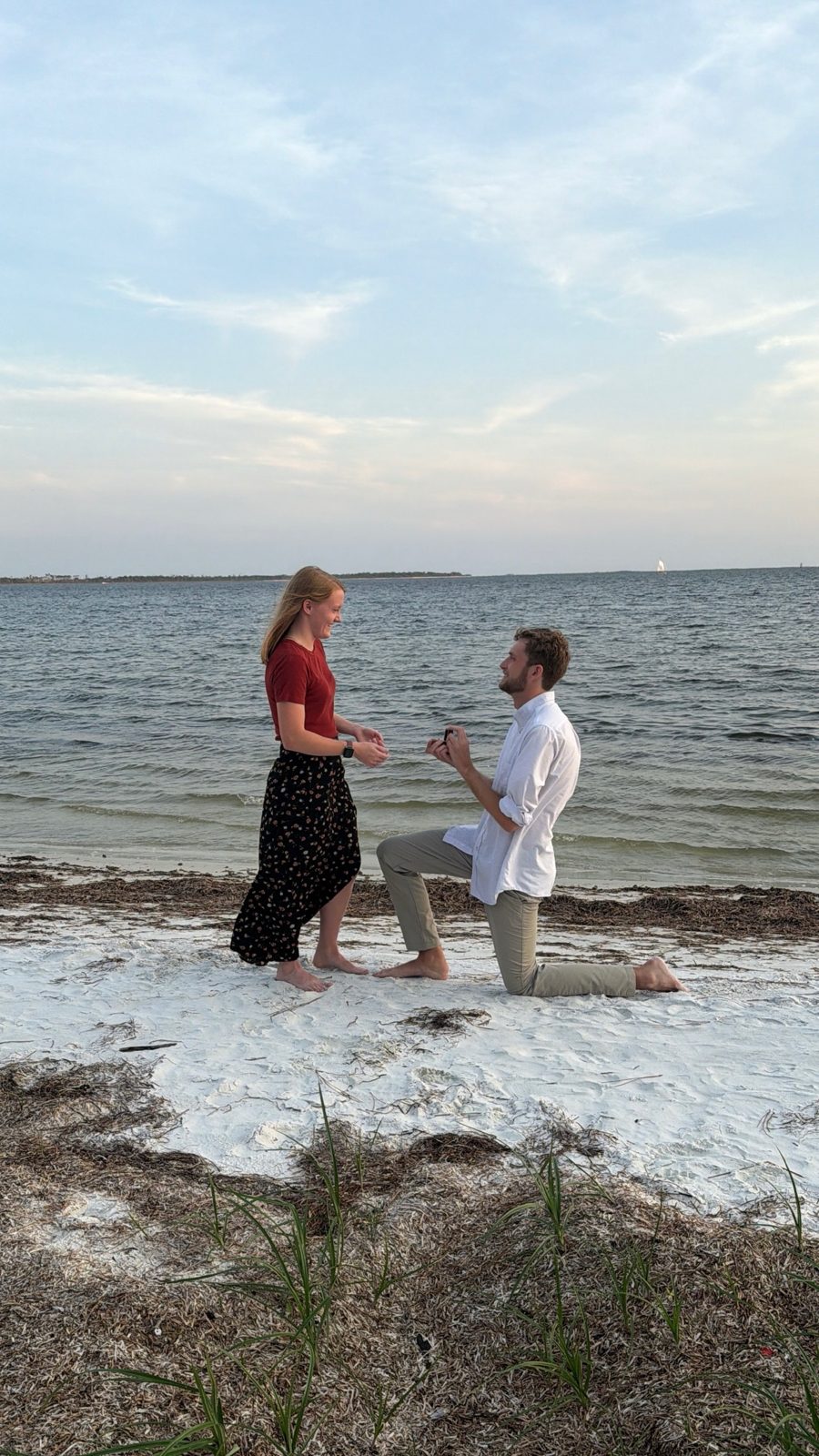 Maria smiling as Daniel kneels on a white-sand beach at sunset to propose